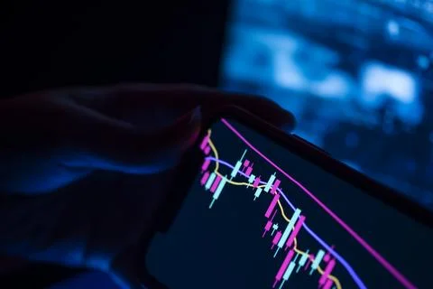 Hand holding a smartphone with a candlestick chart in a dark room Stock Photos