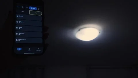 Hand holding smartphone displaying smart lighting options in a dimly lit room Stock Footage 332578523