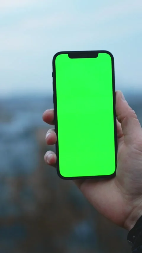 A hand holding a smartphone featuring a green screen while being outdoors in Stock Footage 314151420
