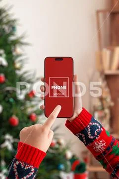 Hand holding smartphone vertically mockup near Christmas tree PSD Template