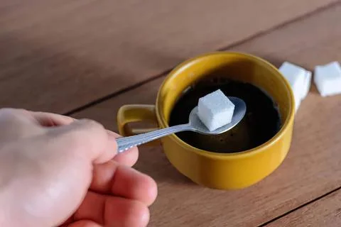 Hand holding spoon with sugar cube over black coffee in mug Stock Photos