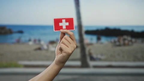 Hand holding swiss flag patch by seaside beach with blurred people in backg.. Stock Footage 306405386