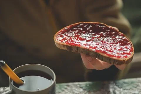 Hand holding toasted dark bread spread with bright red fruit jam beside a ste Фото