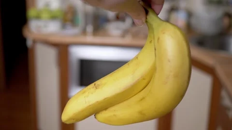 Hand Holding Two Ripe Yellow Bananas in a Blurred Kitchen Setting Stock Footage 339069264