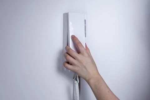 Hand holding a white intercom handset mounted on a wall, minimalist interior  Stock Photos