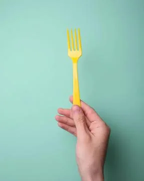 Hand holding a yellow plastic fork against a light green background Stock Photos