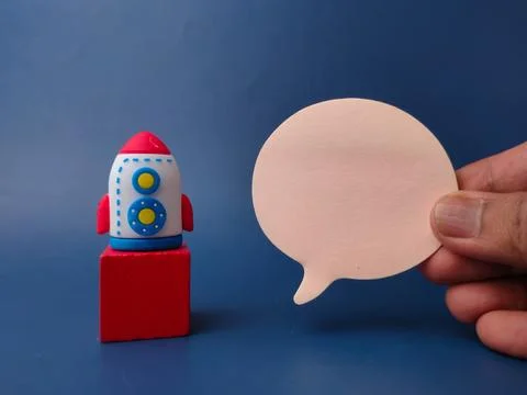 A hand holds a blank speech bubble next to a toy rocket positioned on a red.. Stock Photos