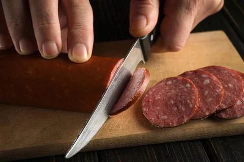 A hand holds a knife to cut thin slices of sausage. The sausage is on a woode Stock Photos