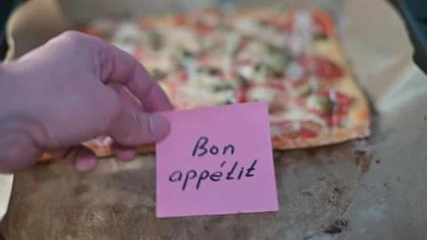 A hand holds a pink note with a message above a fresh pizza on a baking tray. Stock Footage 339096813