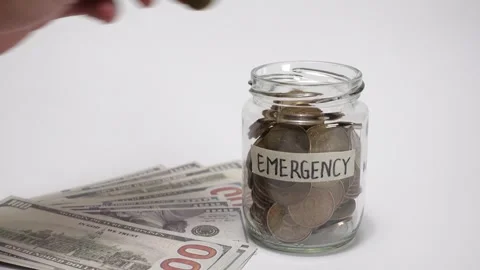 A hand inserting a coin into a labeled emergency savings jar Stock Footage 317108232