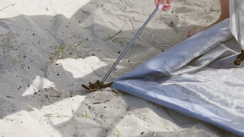 Hand inserting tent pole into loop on sandy beach during daytime camping setu Stock Footage 331200127
