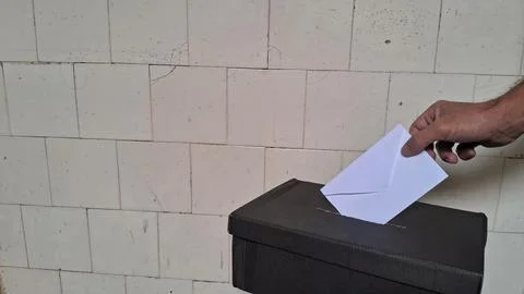 Hand inserting white paper envelope into black cardboard voting box Foto stock