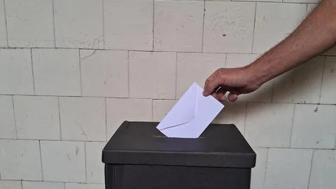 Hand inserting white paper envelope into black cardboard voting box Foto stock