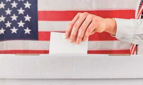 Hand inserts a blank ballot into a box in front of an American flag Stock Photos