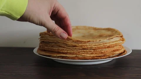 Hand lifting crepe from tall stack of thin golden pancakes on white plate over Stock Footage 309301735