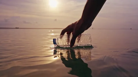 Hand Lifting Crushed Plastic Bottle from Calm Sea at Sunset with Golden Reflecti Stock Footage 329059665