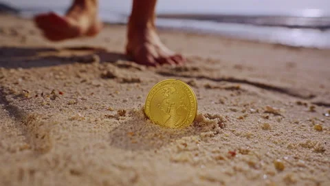 A Hand Lifts a Gold Tether USDT Token From Warm Beach Sand While Soft Sunset Lig Stock Footage 327420133