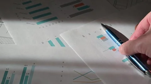 A hand moves a pen over printed business reports that display various graphs and 스톡 동영상 337555826