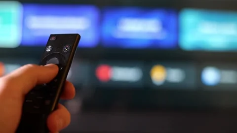 Hand operating a black remote control with a blurred TV background. Stock Footage 340930072