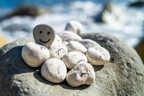 Hand painted smiley face on a small stone. A happy face surrounded by sad faces. Stock Photos