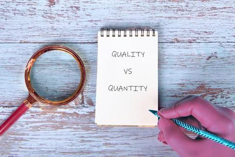 A hand with a pencil points to a notepad displaying QUALITY VS QUANTITY. A ma Stock Photos