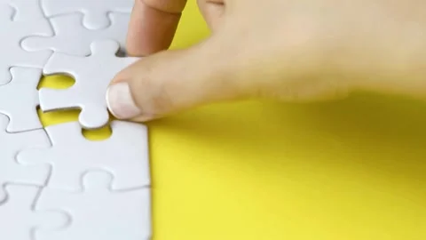 Hand is picking up a white jigsaw puzzle and attaching it to the board Finding a Stock-Footage 336248627