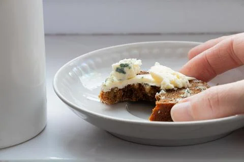A hand picks up a piece of bread and cheese from a plate next to a mug of cof Stock Photos