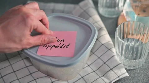 A hand places a note bon appetit on a sealed container with food while glassware Stock-Footage 339096775
