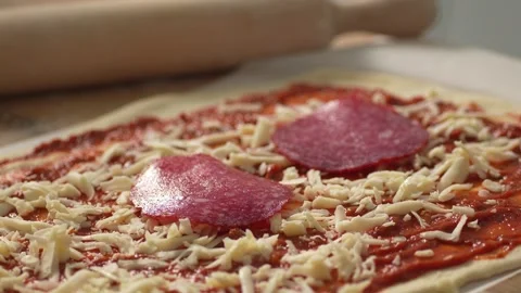 A hand places a round slice of salami on a cheese and tomato-sauced pizza base. Stock Footage 266012089