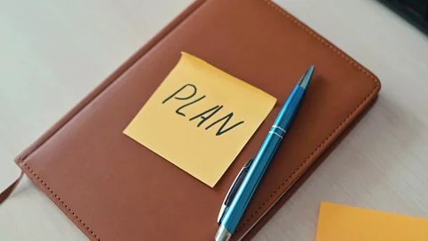 A hand places a sticky note labeled PLAN on a brown notebook. A blue pen lies Stock Footage 339094100