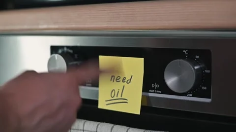A hand places a yellow sticky note on the oven dial as a reminder to buy oil. Stock Footage 339096722