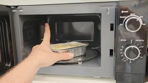 Hand placing cheese-topped meal in foil tray into microwave oven Stock Footage 346412147