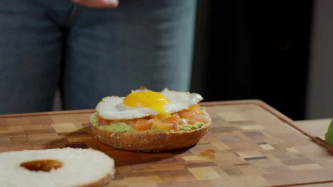 Hand placing fried egg on bagel with salmon and avocado spread Stock Footage 330392927