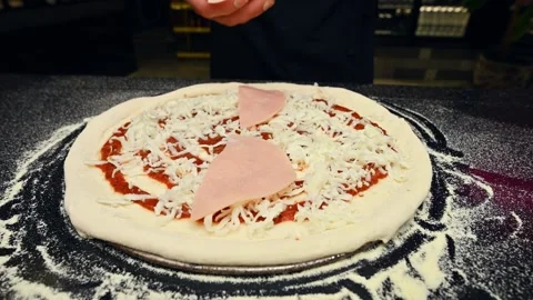 Hand placing ham slices on pizza with cheese and sauce in kitchen setting Stock Footage 337229127