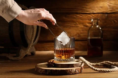 Hand placing large ice cube into whiskey glass with amber liquor on rustic wo Stock Photos