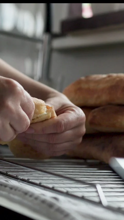 Hand placing split homemade bread roll onto cooling rack beside stacked browned 動画素材 339306730