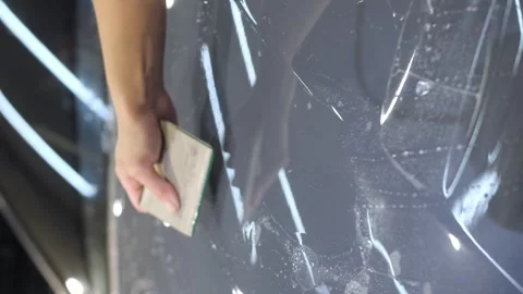 A hand with a plastic scraper glues a protective anti-gravel film on the new car Stock Footage 243803208