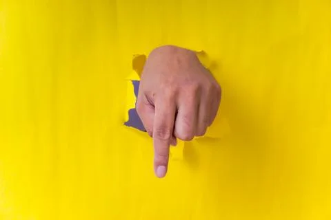 A hand is pointing downward through a torn yellow paper, creating a vibrant.. Stock Photos
