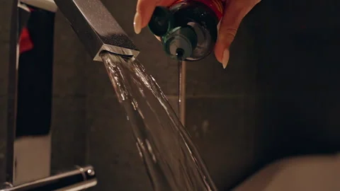 Hand Pouring Bath Foam from Bottle into Flowing Water Under Faucet in a Close An Stock Footage 323469230