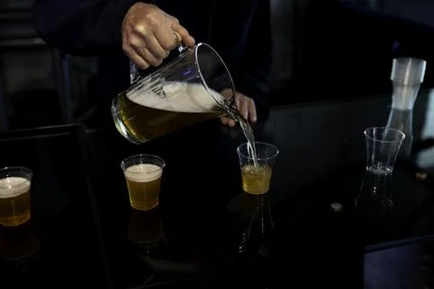 Hand Pouring Beer From Pitcher Into Plastic Cups On Bar Counter At Nighttim.. Stock Photos
