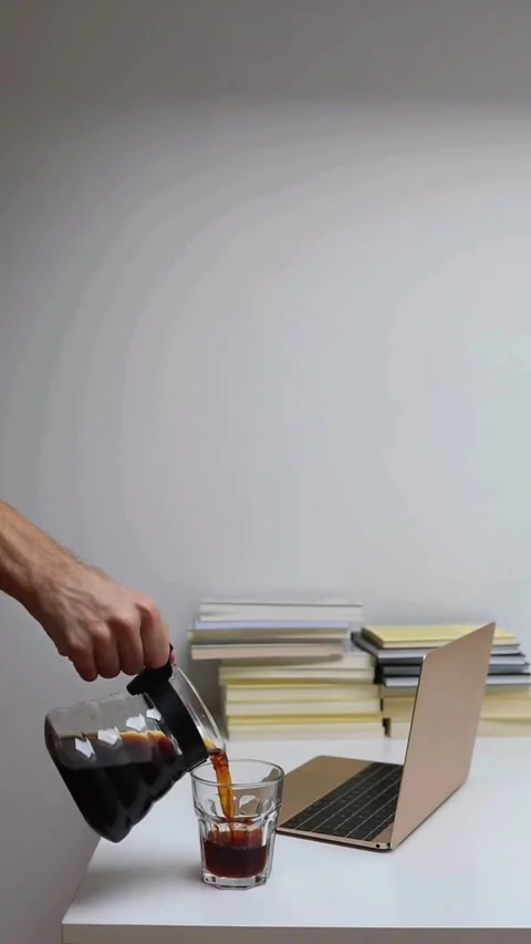 Hand pouring fresh black coffee into a glass cup at a desk with laptop and books Video stock 343471106