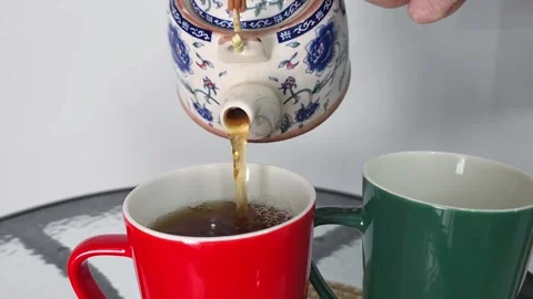 Hand pouring tea from a ceramic teapot into a red mug next to a green mug Stock Footage 282610298