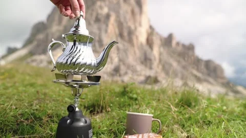 Hand pouring tea from silver teapot on camping stove in alpine meadow with Video stock 319006095