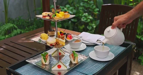 Hand pouring tea from teapot next to three-tier afternoon tea stand Stockbeeldmateriaal 332604899