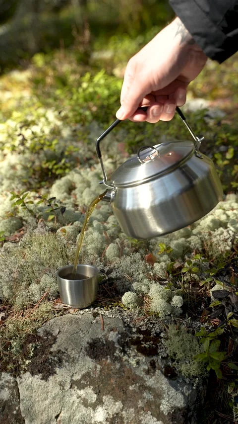 A hand pours hot tea from a stainless steel kettle into a metal cup standing on Video stock 313292552