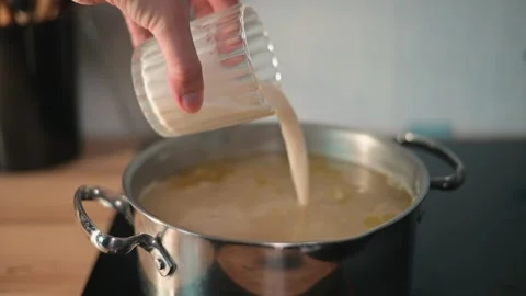 A hand pours a liquid from a container into a pot placed on a stove. The pot Stock-Footage 338301403