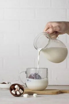 A hand pours milk from a jug into a glass cup with cocoa, chocolate bomb. Making Stock Photos