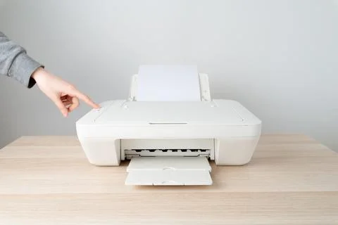 A hand presses the start button on a white home inkjet printer with paper loa Stock Photos