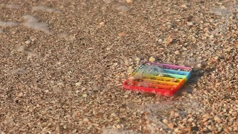 Hand pressing rainbow pop it toy on sand near small ocean wave, Stock Footage 342259629