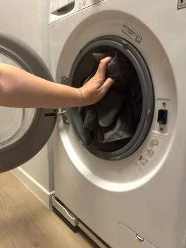 A hand pushes dark laundry inside an open front loading washing machine.  Stock Photos